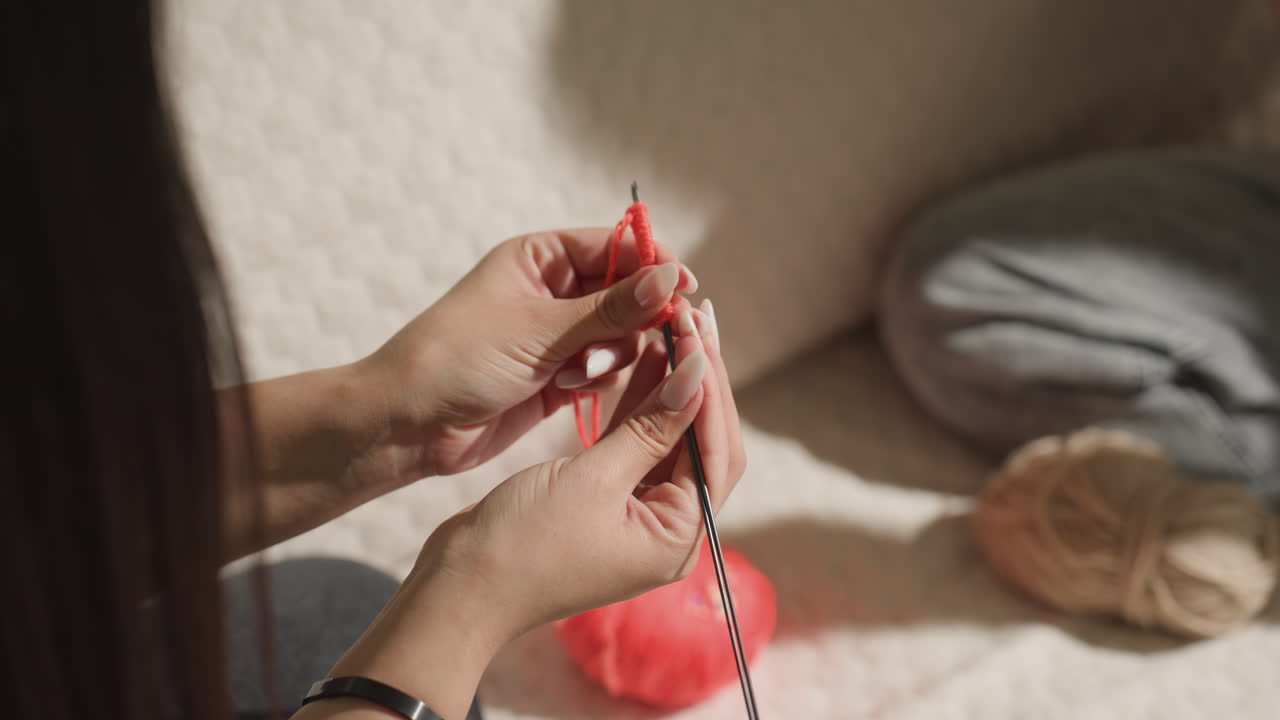 빨간색 털실과 금속 바늘로 뜨개질 기법에 집중하는 백인 여성의 클로즈업, 스티치를 만드는 손가락에 꽉 찬 프레임, 은은한 배경 쿠션, 교육적인 속도와 촉각적 디테일