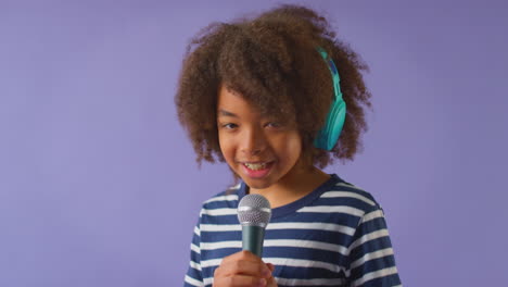 Foto-De-Estudio-De-Un-Niño-Con-Audífonos-Cantando-Karaoke-En-El-Micrófono-Contra-Un-Fondo-Morado