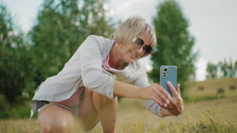 middle-aged lady in sunglasses squatting while taking a selfie with a warm smile in an open field with trees in blurred background, natural sunlight highlights her joyful expression