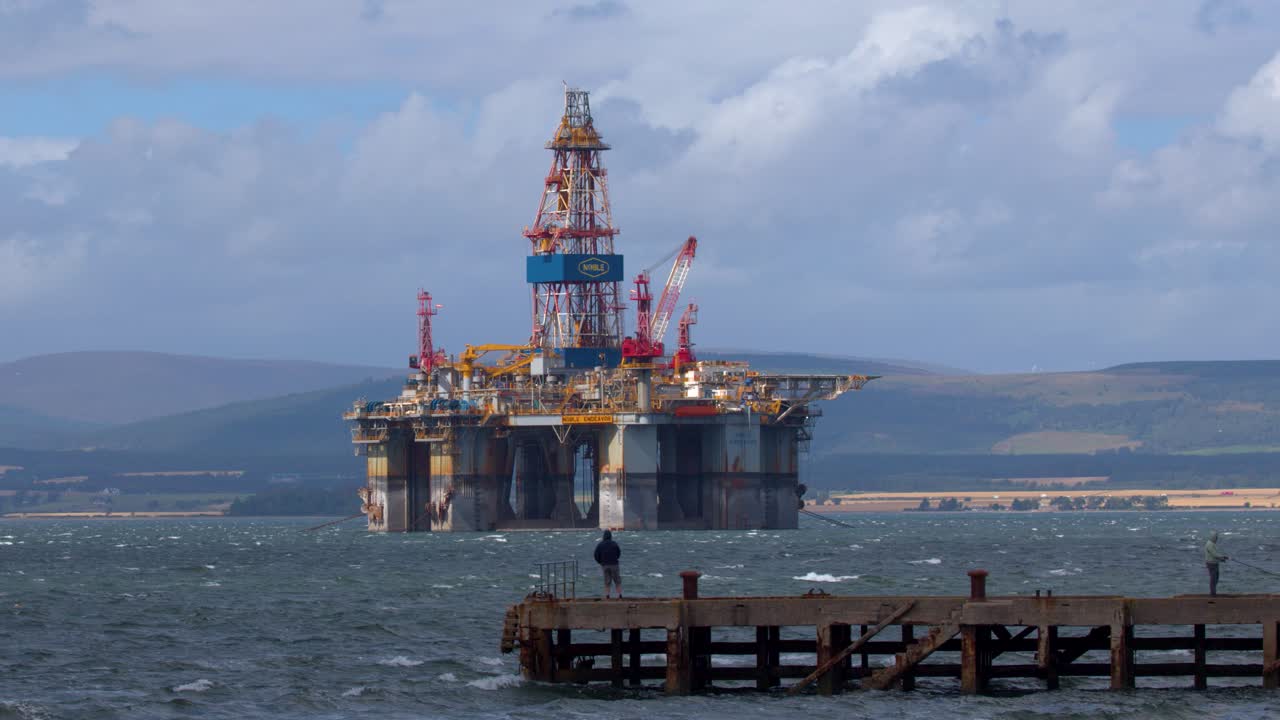 Stor offshore-olieplatform bevæger sig mod træmole, fisker stående, dagslys, roligt kamera, Skotlands kyst