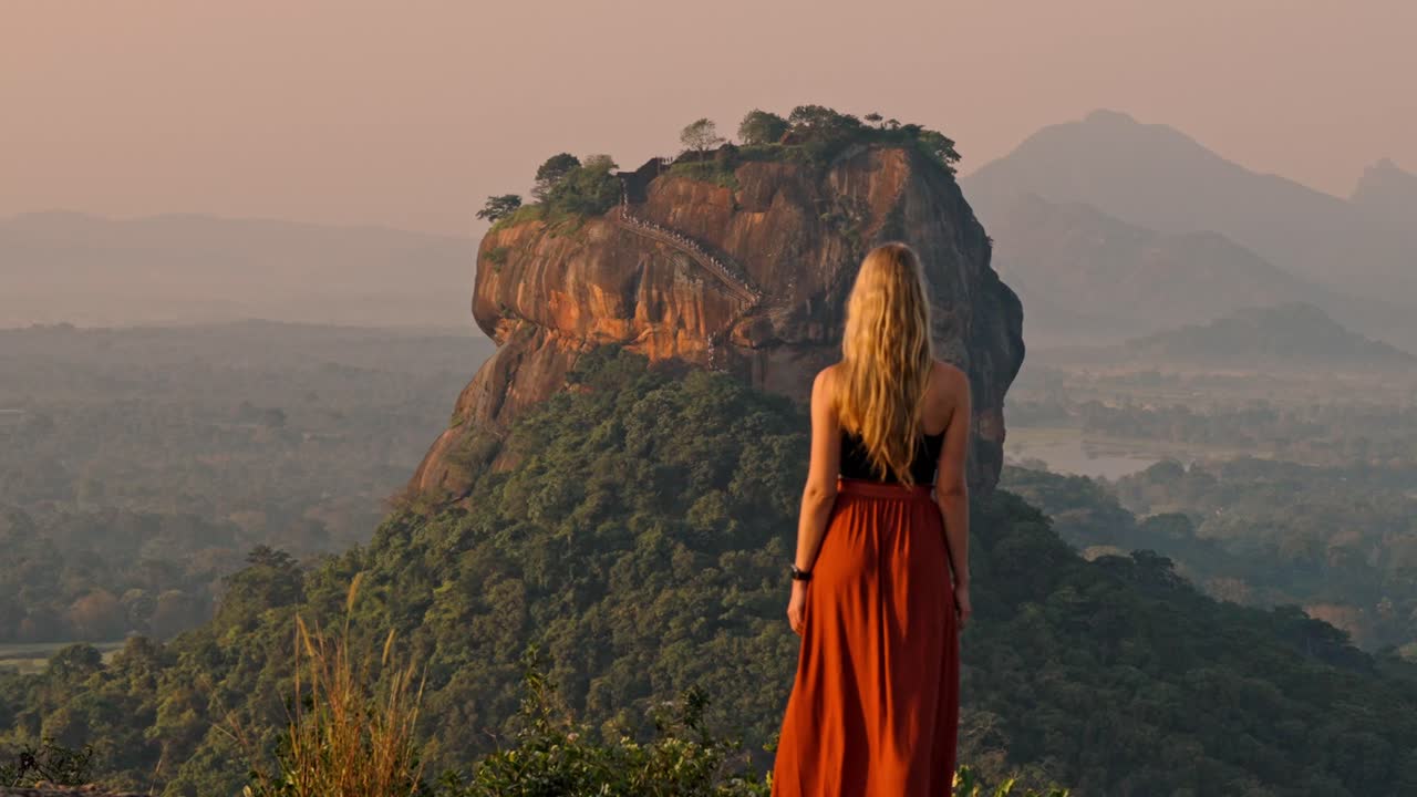 A lone woman stands amid lush greenery, gazing toward the ancient Sigiriya Rock fortress as the sun rises over Sri Lanka.