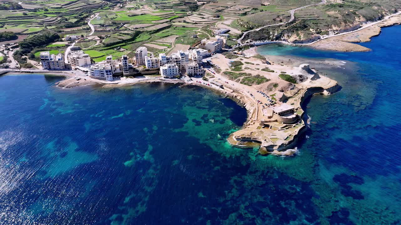 Upea panoraamallinen korkeakuvakulman dronekuva Gozon saaren rannikkokylästä kirkkaana kesäpäivänä, esitellen eloisia sinisiä turkooseja vesiä, jylhiä kalkkikivikallioita ja vehreitä terassoituja viljelysmaita.
