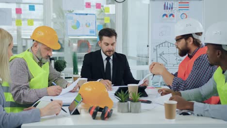 handsome skilled bearded head chief discussing with his multiracial subordinates-builders or architects the details of building plan in boardroom