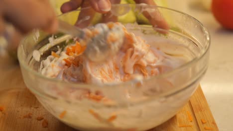 close-up of hands mixing grated carrots and creamy sauce in a glass bowl
