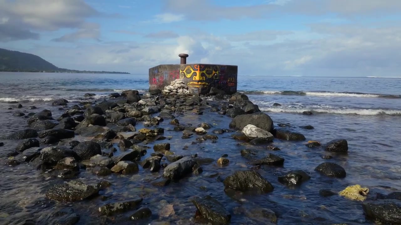 Gadekunst ved havet, Taiarapu-vejen til Teahupoo, Tahiti, Fransk Polynesien. Droneoptagelse af Stillehavet, cementblokmole, graffiti-maleri af et polynesisk ansigt, lavvandssten, bølger der slår ind, panorering op.