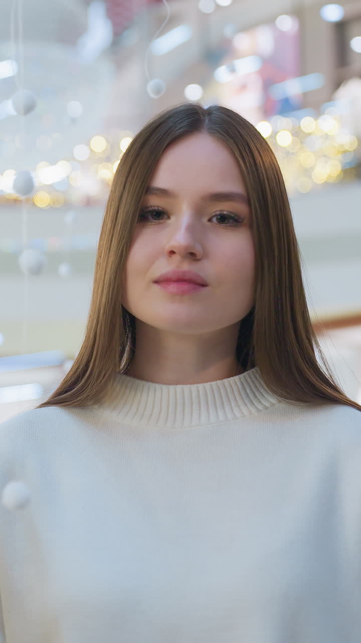 Elegant lady in white sweater standing near beautifully decorated Christmas tree in festive mall setting, surrounded by ornaments, twinkling lights, with blurred shoppers in background