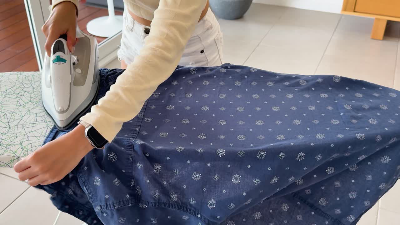 A young woman uses a steam iron to press a blue patterned shirt on an ironing board in a bright, modern indoor setting with natural light