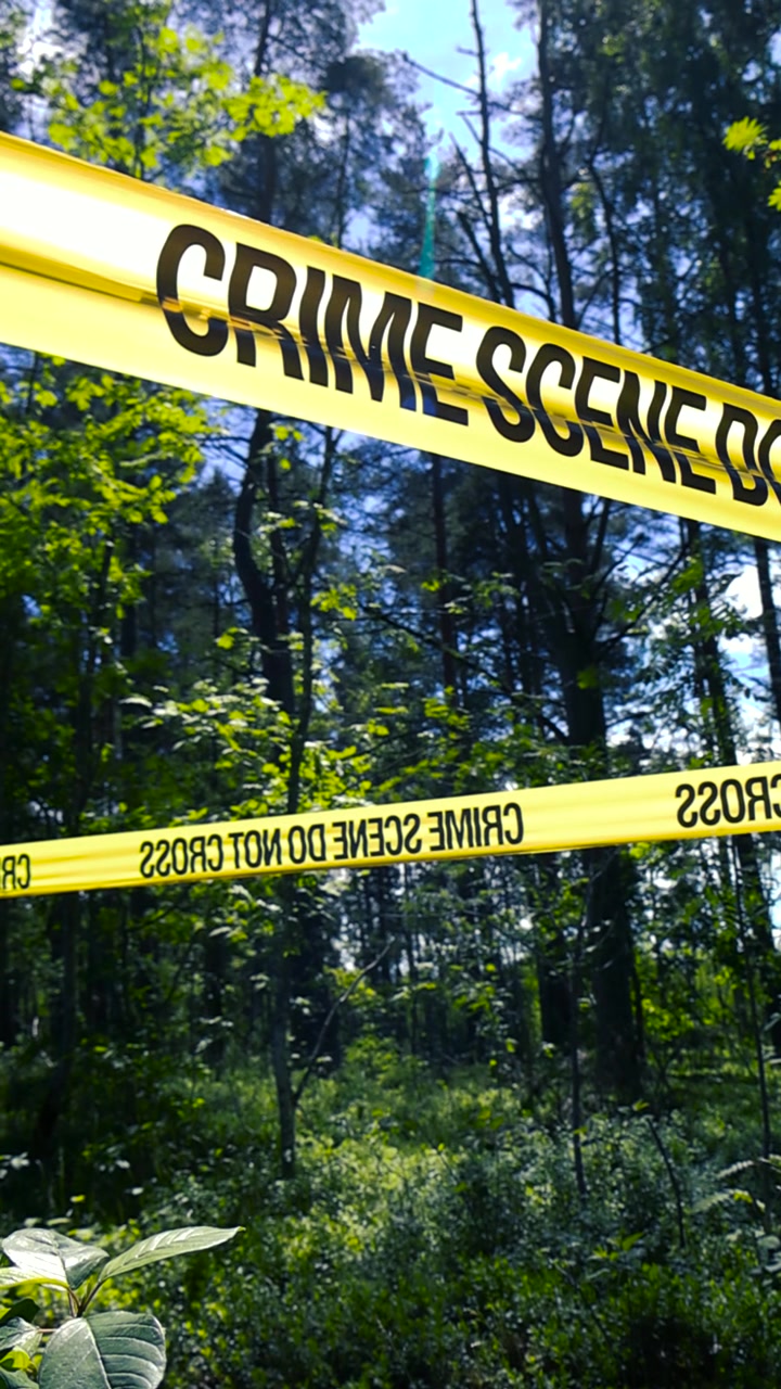 Yellow police tape with the words crime scene do not cross cordoning off a section of a lush green forest, signaling an ongoing investigation and restricting access to the area