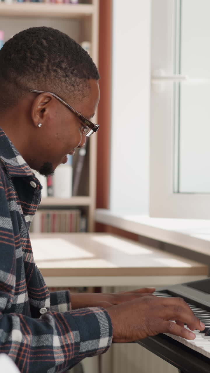 un hombre presiona las teclas del sintetizador por la estantería. un músico afroamericano sonriente practica la afinación en un dispositivo digital en clase. lecciones domésticas de música