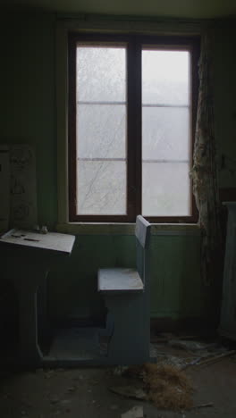 vertical shot of an old wooden school desk and bench sit in an abandoned classroom with a cracked window and peeling paint.