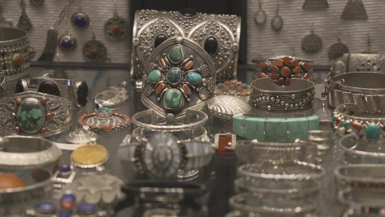 Close-up view of a group of traditional silver jewelry displayed on a glass shelf. The shot features intricate bracelets and rings set with turquoise and coral stones in a Moroccan souk