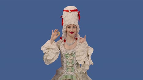 retrato de una dama de la corte con un vestido vintage y una peluca que muestra una moneda de bitcoin y hace un gesto de pulgar hacia arriba. mujer joven posando en el estudio con fondo de pantalla azul. primer plano. cámara lenta lista 59.94fps
