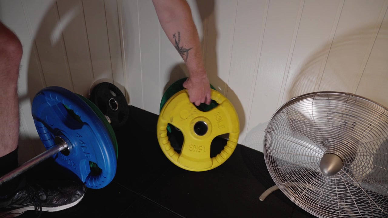 Záběr z vysokého úhlu na roh domácí posilovny s olympijskými vzpěračskými kotouči a běžícím ventilátorem. Mužská ruka zvedá a odstraňuje 15kg žlutý kotouč