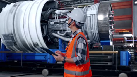 side view of asian male engineer with safety helmet looking at the tablet in his hand and looking around while standing with airplane engine maintenance conducted, analytics checking the turbine