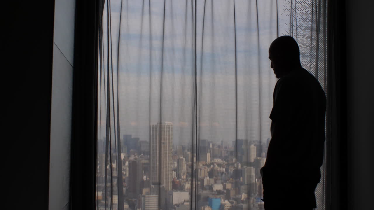 한 남자의 실루이 앞뒤로 걸어가 닫힌 커튼이 있는 창문 밖을 쳐다보며 광활한 도시 대도시 전망을 감추고 있다.