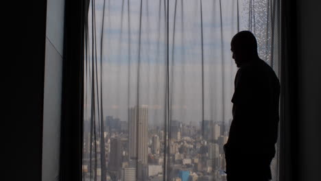 a man's silhouette paces back and forth, gazing out a window with closed curtains, concealing the expansive city metropolis view