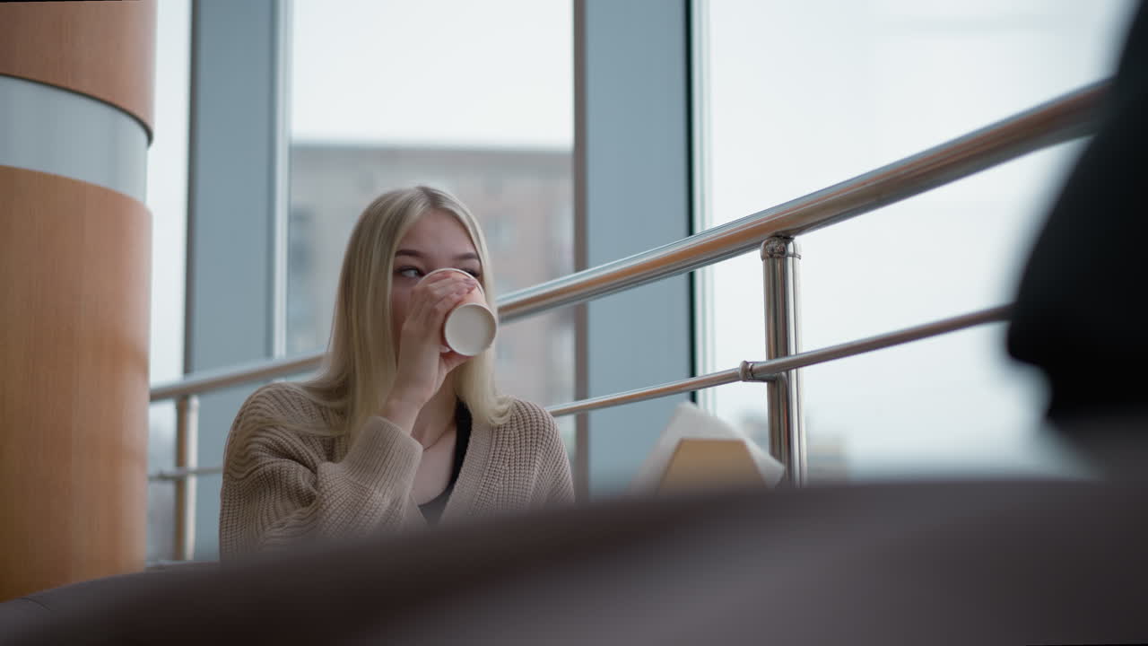 Jovem mulher tomando chá de sua xícara, derrubando-a sobre a mesa enquanto olha para fora através de um vidro transparente em um shopping com luz suave filtrando-se pelo vidro.