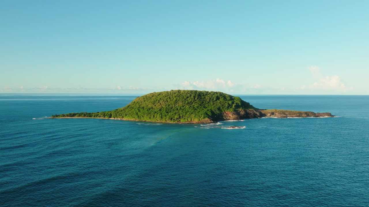 Drone flies forward and ascends slightly toward a small uninhabited island covered in dense vegetation under clear blue sky