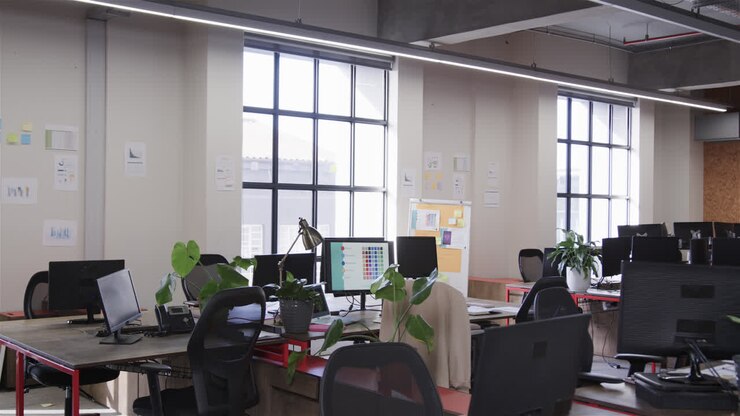 Espacio de trabajo de oficina con grandes ventanales, sillas y escritorios con ordenadores, lámparas y plantas, cámara lenta