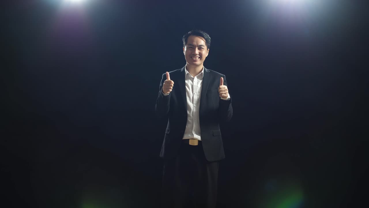블랙 스크린 스튜디오에 서 있는 동안 카메라에 엄지손가락을 올리는 웃는 아시아 스피커 남성