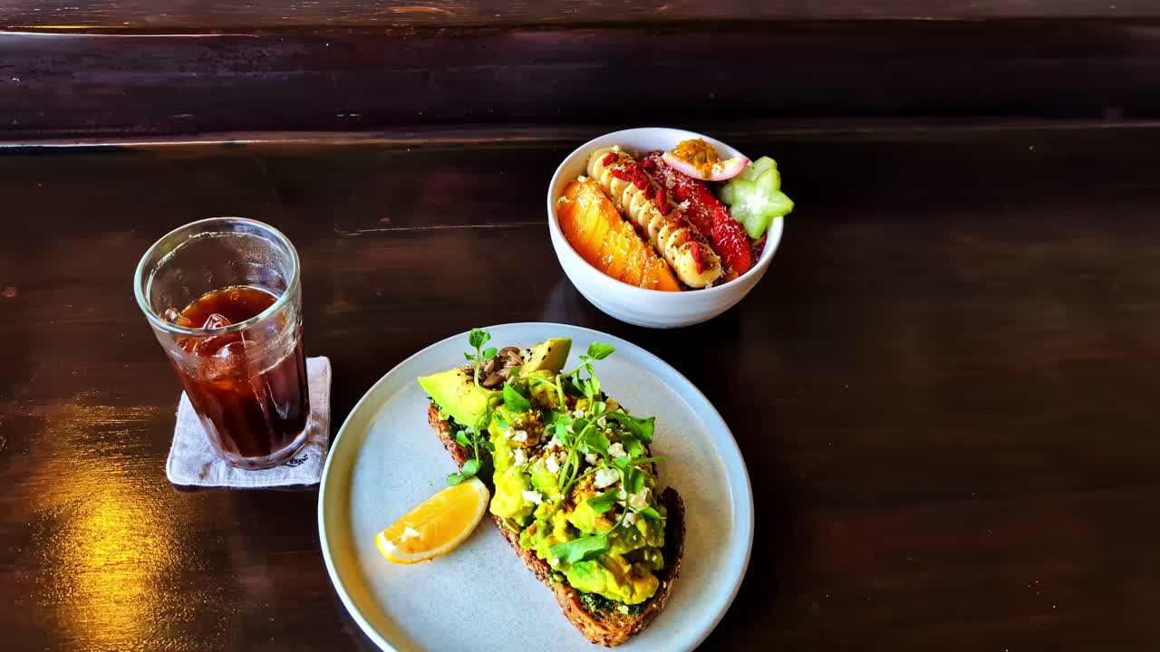 Close-up of a vibrant Bali brunch featuring avocado toast, a tropical fruit smoothie bowl, and iced coffee on a wooden table in Ubud, slow push-in shot