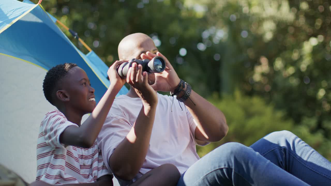 Un padre africano e il figlio adolescente sollevano i binocoli, guidando e osservando gli uccelli in un accampamento vicino a una tenda blu.