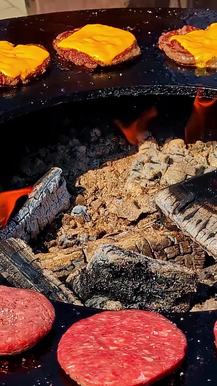 Primo piano di hamburger di manzo e formaggio fuso che cuociono su una griglia metallica circolare sopra ceppi di legno ardenti e braci calde. Autentica scena di barbecue estivo all'aperto.