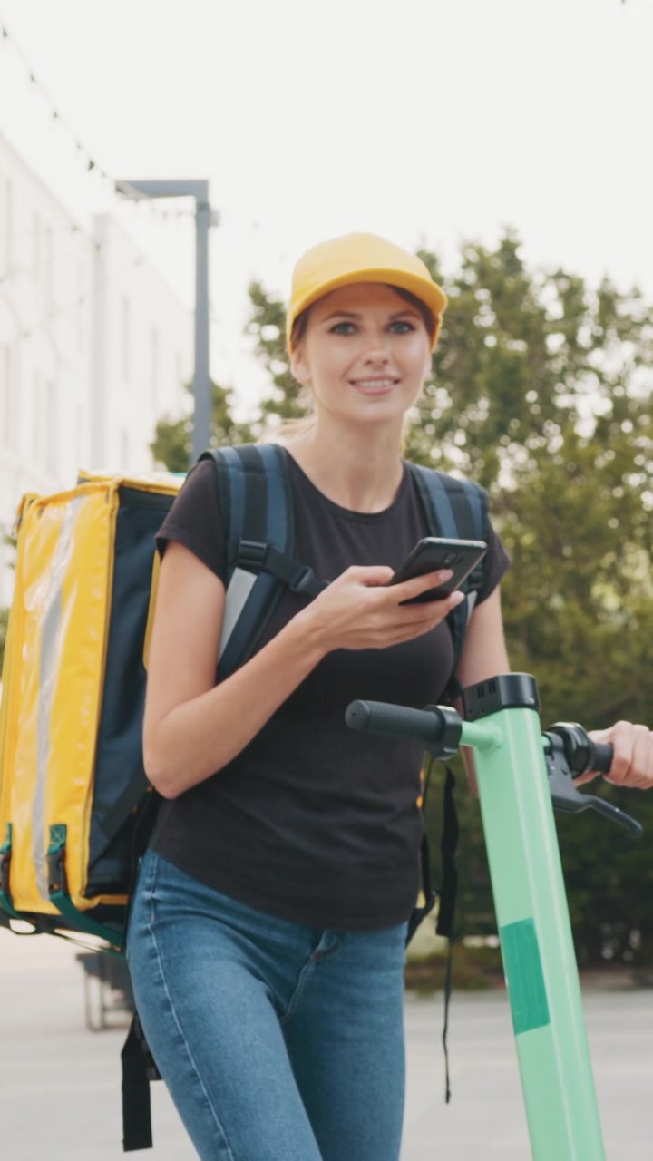 A delivery woman checks her phone while standing next to her scooter ready to make a delivery