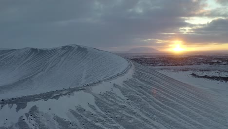 Toma-Aérea-Hacia-Atrás-Del-Cráter-Del-Volcán-Hverfjall-Cubierto-De-Nieve-Durante-La-Puesta-De-Sol-En-El-Horizonte---Norte-De-Islandia,-Europa