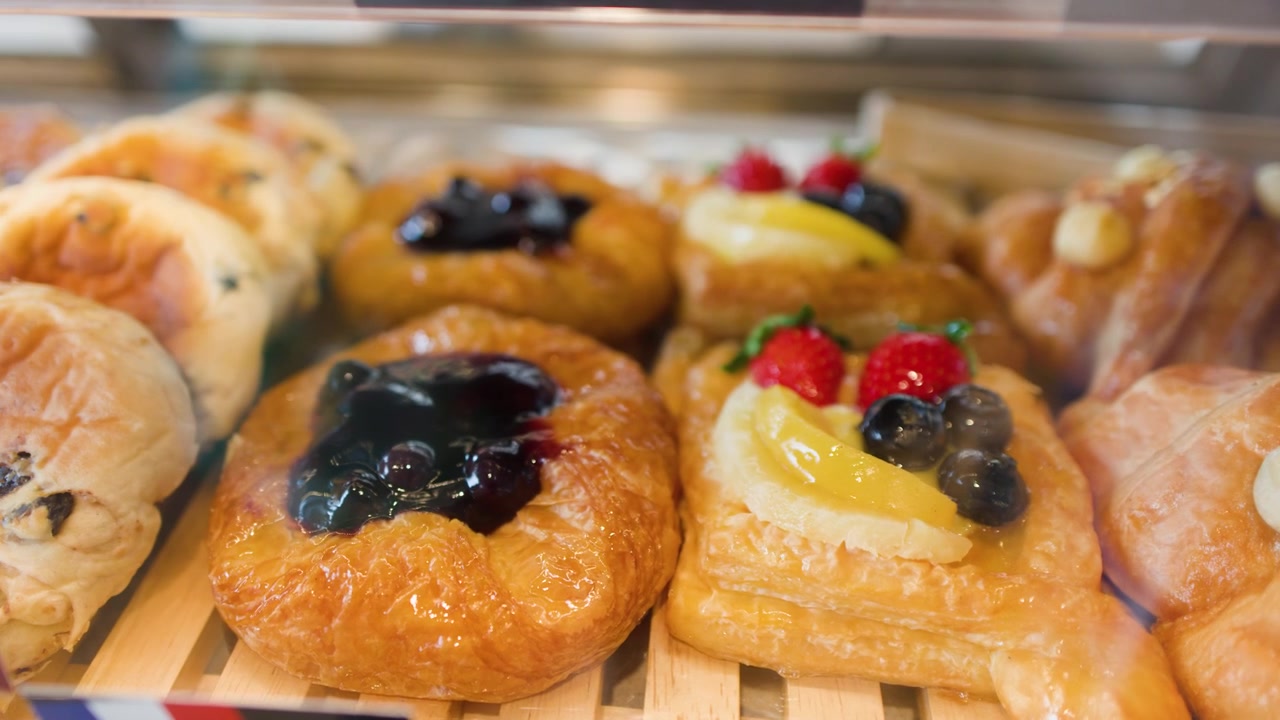 Slow camera pan across a glass display case filled with fresh fruit danishes and buns
