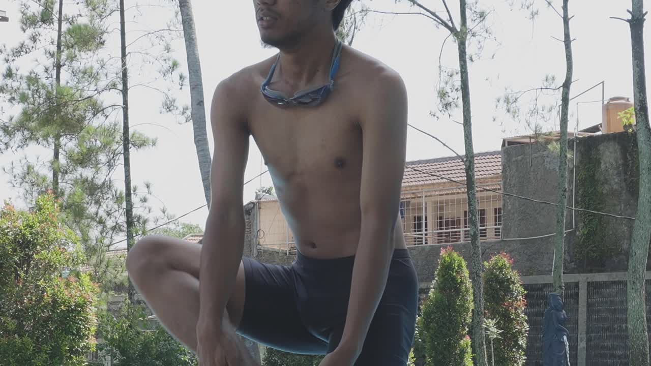 joven asiático lleva pantalones negros sostiene las piernas y las rodillas haciendo calentamiento antes de nadar en la piscina