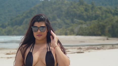 Hispanic-girl-passes-her-hand-in-her-hair-while-sitting-at-the-beach-with-mountains-and-the-ocean-in-the-background