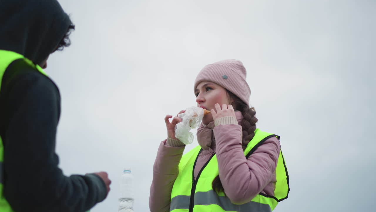 Young woman in pink winter clothing and reflective safety vest eating sandwich outdoors during cold cloudy day, standing next to coworker in safety gear, looking attentive and thoughtful