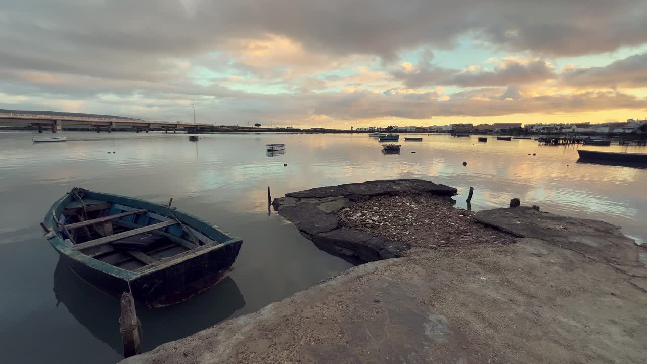 스페인 안달루시아의 바르바테에 있는 붕괴된 콘크리트 도크에 정박한 오래된 목조 시 배가 해가 지는 동안 조용한 물과 다채로운 반사에 따뜻한 빛을 아니다.