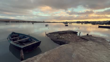 old wooden fishing boat moored by a crumbling concrete dock in barbate, andalusia, spain, as the sun sets, casting a warm glow on tranquil waters and colorful reflections