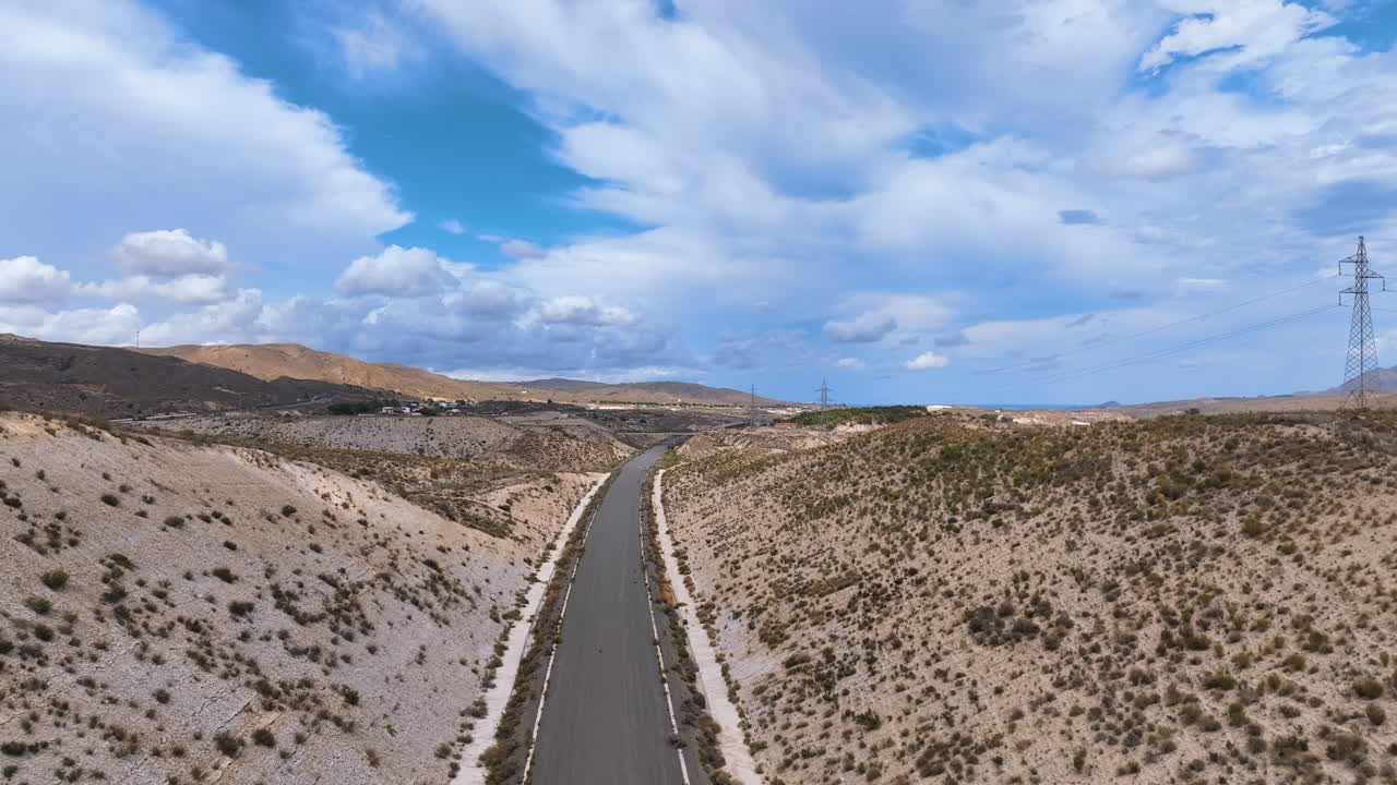 Aerial glides over an emblematic landscape of tussock spotted hills and a long grey vacant road, slowly passing electricity pylons Andalusia Spain.
