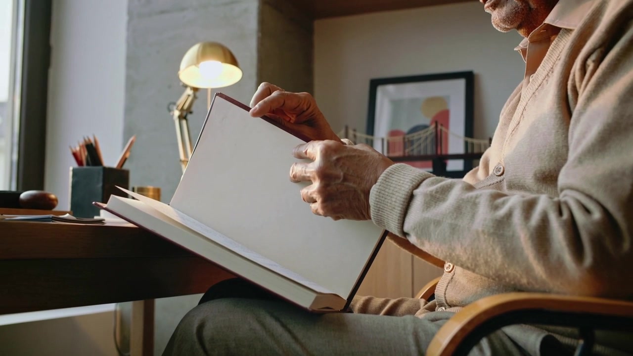 Uomo anziano con maglione beige che inclina un libro, sfoglia le pagine e ispeziona uno sketchbook nello studio di casa, con una lampada.