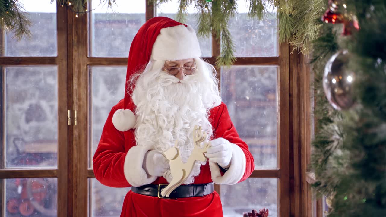 Santa Claus standing in specially decorated room, holding wooden dear near the New year tree. Christmas spirit concept close up portrait. Christmas eve. New Year is coming.