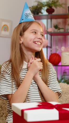 child girl kid celebrates birthday happily opens gift with excited surprise at table on home sofa
