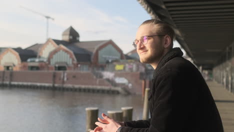 Junger-Europäischer-Mann-Mit-Brille,-Bart,-Manbrötchen-Und-Schwarzer-Kleidung-Genießt-Die-Sonnige-Aussicht-Von-Der-Brücke-In-Hamburg,-Deutschland