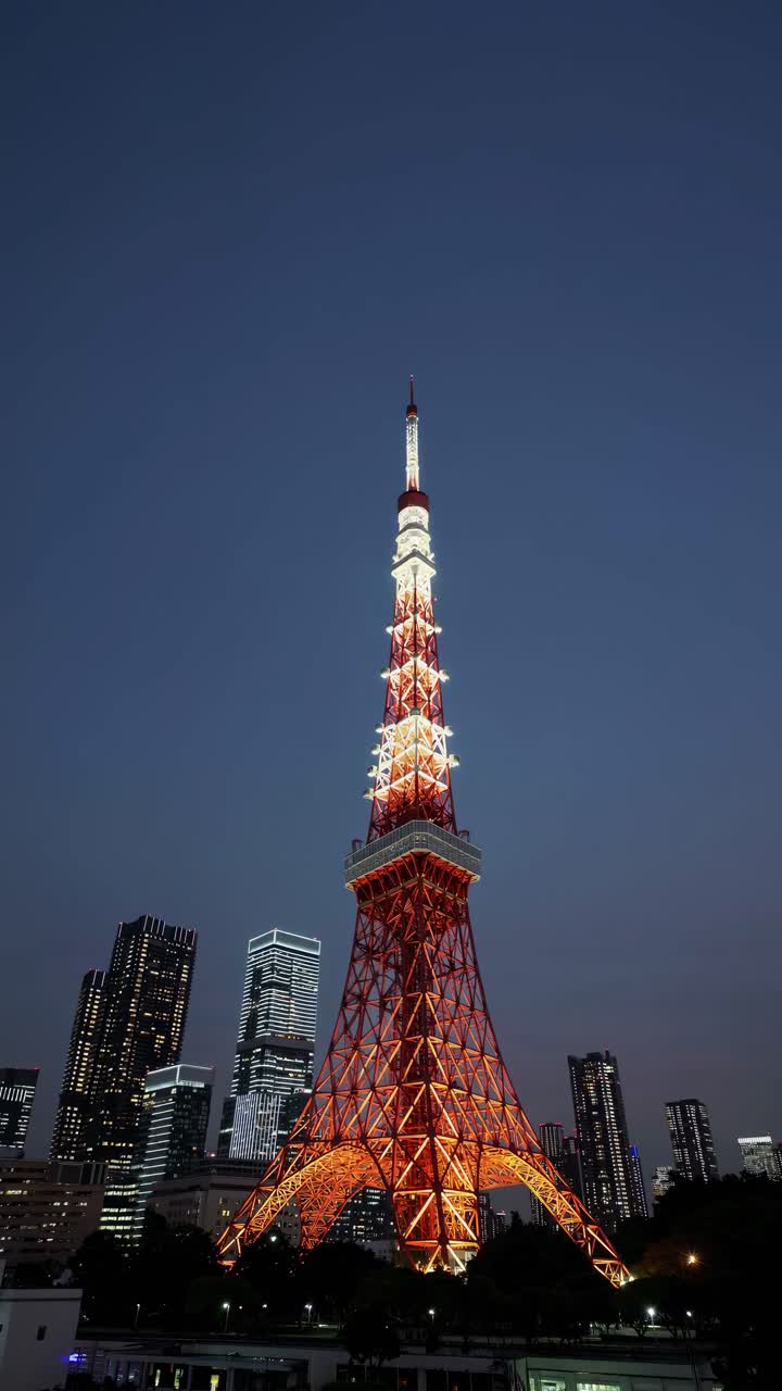 A low-angle shot of a brightly lit tower against a twilight sky, surrounded by city skyscrapers