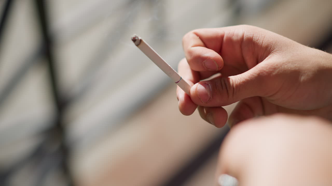 close up hand view of smoker gently flicking cigarette as ash falls with subtle smoke trail against softly blurred outdoor setting in daylight