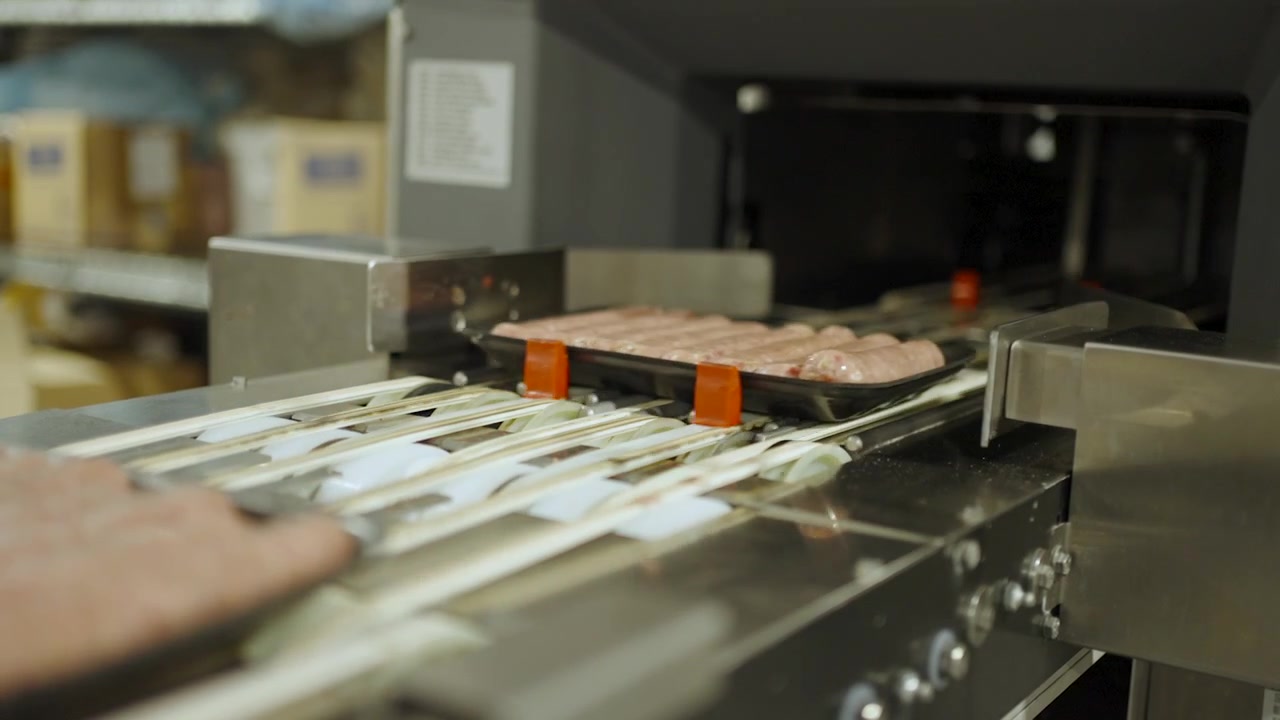 Tray-packed sausages travel along a stainless conveyor into an inspection scanner inside a food factory.