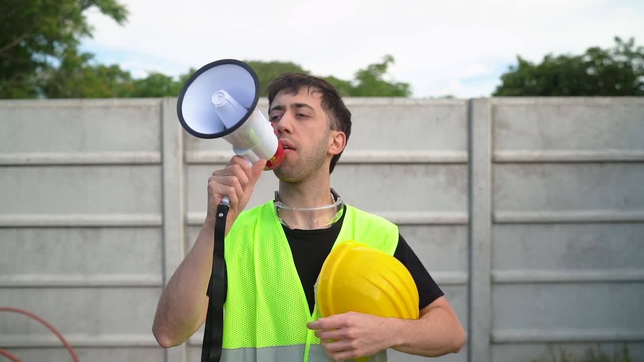 un trabajador de la construcción, agarrando su casco amarillo, emplea un megáfono para comunicarse con el equipo - medio de primer plano