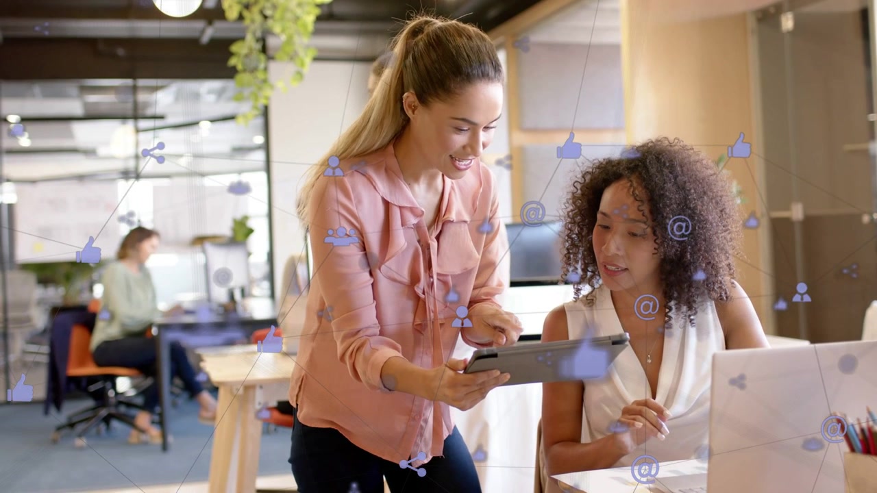 Standing woman presenting tablet to seated coworker at laptop for feedback, tech icons drifting