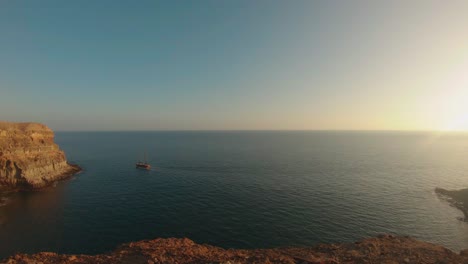 Puesta-De-Sol-En-La-Playa-De-Tiritaña,-Gran-Canaria,-España,-Con-Un-Barco-Navegando-Y-Un-Cielo-Azul-Enmarcado-Por-Altos-Acantilados-Cerca-De-Taurito-mogas,-Entre-Punta-De-La-Cruz-De-Piedra-Y-Punta-De-Los-Medios-Almudes