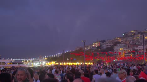 a large crowd gathers in the city at night, under a dark sky, with a bridge in the background.