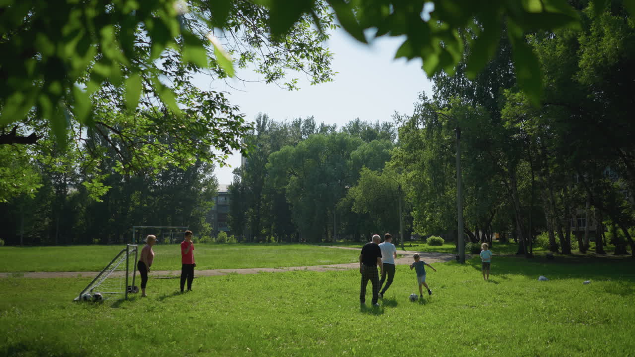 Family enjoys an outdoor soccer game on a sunny day as a little boy plays close to his grandfather, who moves back, and the boy kicks the ball towards the goal post
