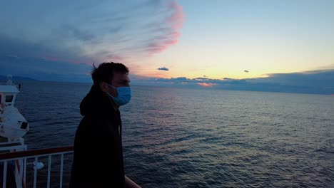 side view of a man with dark brown hair and a blue jacket with mask, looking at clouds from ferry boat which is sailing from split to island vis in croatia, adriatic sea. during a corona pandemic.