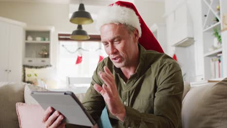 Glücklicher-Kaukasischer-Mann-Mit-Weihnachtsmannmütze-Und-Tablet-Für-Videoanrufe-Im-Wohnzimmer